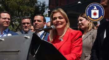 Primer plano de una mujer rubia con blazer rojo hablando en un micrófono. Detrás, personas y el logo de la CIA en la esquina superior derecha.
