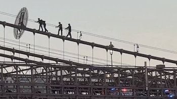 Siluetas de tres personas sobre un cable de puente, una lleva objeto circular. Vehículos policiales con luces en el nivel inferior