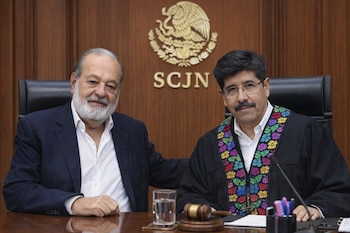 Dos hombres sonriendo sentados ante un fondo de madera con el logo de la SCJN. Uno viste traje y camisa blanca, el otro una toga negra con bordados de flores de colores.