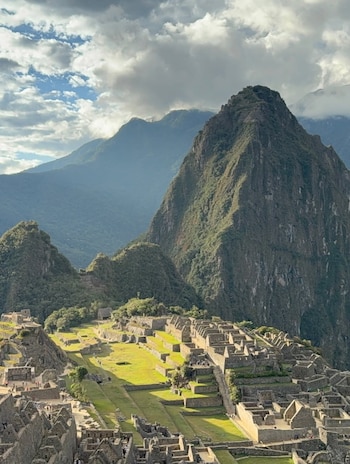 Vista aérea de la ciudadela inca de Machu Picchu con estructuras de piedra, terrazas verdes y montañas, bajo un cielo con nubes grises y zonas azules