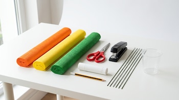 Rollos de papel crepé, tijeras rojas, abrochadora negra, adhesivo, lápiz, alambres verdes y un vaso plástico transparente sobre una mesa blanca.