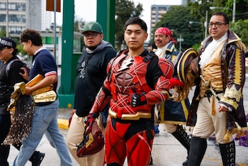 Un hombre joven con un traje de superhéroe rojo y negro con símbolo de rayo y un casco en la mano, camina junto a otras personas, algunas disfrazadas
