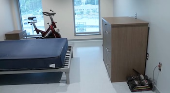 Interior de habitación con cama de colchón azul, mesita de noche, bicicleta estática roja, cómoda de madera clara y pesas en el suelo. Ventanas grandes