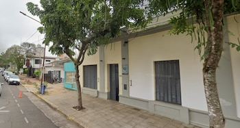 Exterior de un edificio color crema con detalles grises, una puerta y dos ventanas con persianas. Una calle con automóviles, árboles y acera