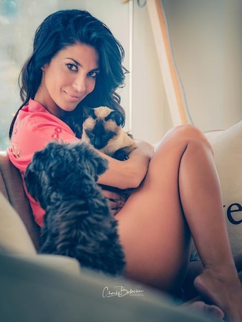 Silvina Escudero sonriendo a cámara, sentada con las piernas cruzadas, sosteniendo un gato siamés y un perro de pelaje oscuro. Lleva una bata rosa