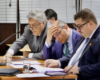 Tres hombres con traje y anteojos leen documentos en una mesa equipada con micrófonos y vasos de agua