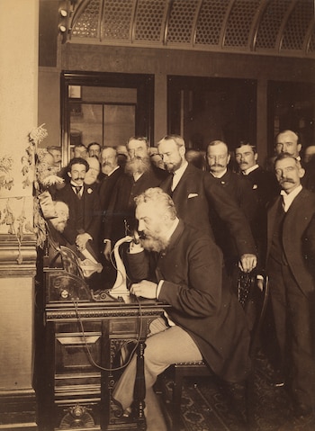 Fotografía sepia de Alexander Graham Bell, un hombre barbudo, sentado y hablando por un teléfono antiguo rodeado de varios hombres observando