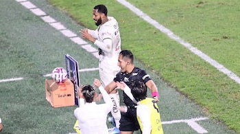 Un árbitro vestido de negro y con auricular habla con tres futbolistas vestidos de blanco junto a la línea de la cancha.