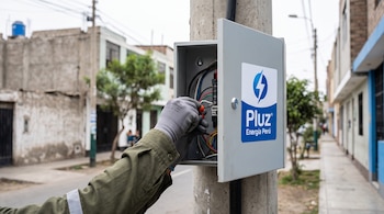 Mano con guante maniobra cables con un destornillador dentro de una caja metálica de electricidad en un poste con el logo de Pluz Energía Perú.