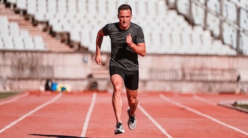 Un hombre corre a alta velocidad sobre una superficie deportiva al aire libre, con el cuerpo ligeramente inclinado hacia adelante, los brazos flexionados y una zancada amplia, en una postura que refleja potencia, coordinación y agilidad durante el entrenamiento.