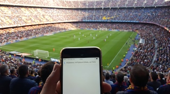 Primer plano de un smartphone en una mano con 'ROAMING INTERNACIONAL ACTIVO' en pantalla, y al fondo, un partido de fútbol en un estadio lleno.