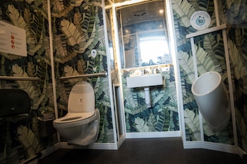 Un baño con inodoro, urinario, lavabo y espejo, sobre un papel tapiz con estampado de hojas verdes