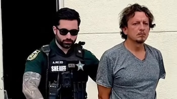 Un oficial de policía con gafas de sol y uniforme verde escolta a un hombre con camiseta gris y cabello desordenado. Fondo de pared clara