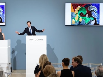 Hombre en traje gesticulando detrás de podio blanco con logo Phillips. Dos pantallas muestran obras de arte abstractas. Mujer y público sentados
