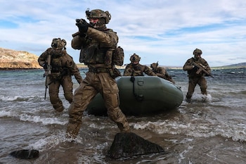 Cinco soldados con uniforme militar de camuflaje y cascos desembarcan con una barca inflable sosteniendo fusiles en una playa