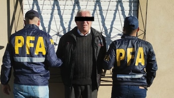 Hombre de edad avanzada, con el rostro censurado, flanqueado por dos agentes de la PFA con uniformes azules y letras amarillas, frente a una persiana metálica