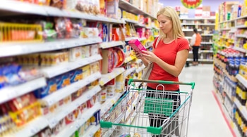Alergias alimentarias: qué exigen las nuevas reglas para el etiquetado de productos y su impacto en la Argentina 7 Mujer rubia con camiseta roja sostiene una caja rosa, al lado de un carro de compras verde, en un pasillo de supermercado con estantes de productos.