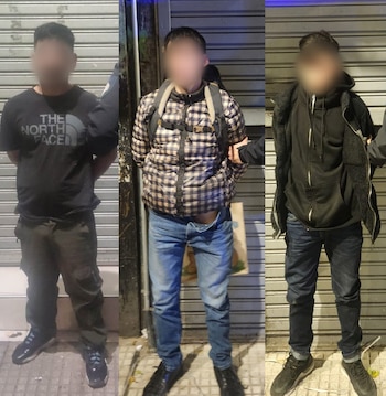 Tres hombres con rostros difuminados, de pie. Uno con camiseta negra. Uno con chaqueta a cuadros y bragueta abierta. Uno con sudadera negra. Manos en la espalda. Fondo de persianas metálicas