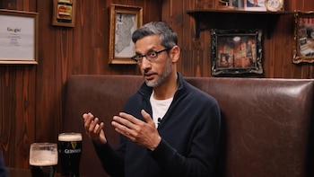 Sundar Pichai, con gafas y una chaqueta azul oscuro, sentado en un sillón de cuero marrón. Gesticula con las manos cerca de dos vasos de cerveza oscura