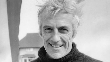 Retrato en blanco y negro de un hombre mayor con pelo revuelto, cuello alto y una amplia sonrisa. Su rostro tiene arrugas