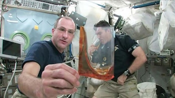 Dos hombres en el interior de una nave espacial sostienen recipientes plásticos transparentes con un líquido de tono marrón