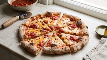 Una pizza redonda cortada en ocho porciones sobre una mesa de mármol clara. Hay un cortador de pizza, un cuenco con salsa de tomate y un recipiente con aceite.