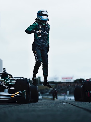 Un piloto de carreras con traje verde y casco azul salta en el aire sobre una pista. Se ven partes de dos coches de carreras a ambos lados del piloto