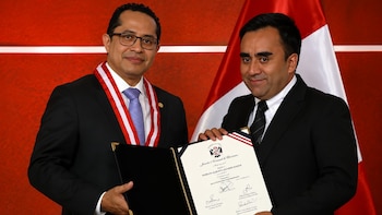 Dos hombres vestidos de traje sostienen una credencial enmarcada del Jurado Nacional de Elecciones ante una bandera de Perú y un fondo rojo