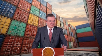 Un hombre con traje y corbata roja, Marcelo Ebrard, habla en un podio con el escudo de México. Detrás hay pilas de contenedores de carga.