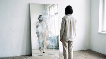 Persona de espaldas con cabello oscuro y ropa clara. Un espejo rectangular grande muestra un reflejo borroso de una figura. Pared blanca y suelo de madera.