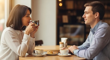 Una mujer y un hombre están sentados en una mesa de café. Ella sostiene una taza plateada y la mira, mientras él la observa. Hay tazas con café y dulces en la mesa.