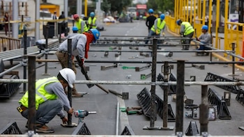 Varios trabajadores con cascos y chalecos reflectantes instalan estructuras metálicas en una avenida. Hay otros trabajadores y vehículos al fondo