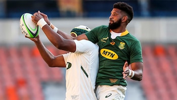 Dos jugadores de rugby, uno con camiseta verde y otro con camiseta blanca, extienden las manos hacia un balón ovalado