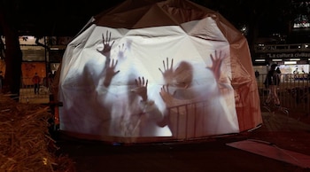 Carpa domo blanca iluminada de noche con siluetas de manos y personas proyectadas sobre la tela en una plaza pública con vallas metálicas