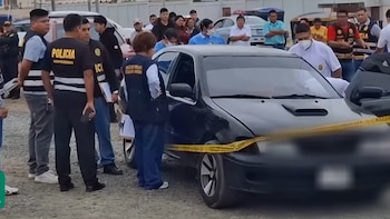 Personas con chalecos policiales y forenses rodean un automóvil negro. Una cinta amarilla de seguridad delimita el vehículo en zona exterior