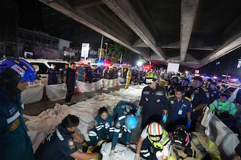 Personal de emergencia, personas tendidas bajo sábanas, vehículos de emergencia con luces y edificios con letreros en tailandés, bajo un paso elevado