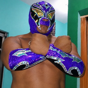Un hombre con el torso descubierto viste una máscara morada brillante y coderas a juego mientras cruza los brazos sobre el pecho