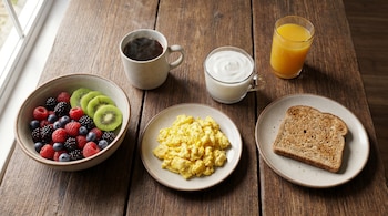 Un desayuno variado sobre una mesa de madera, incluyendo un tazón con bayas y kiwi, huevos revueltos, yogur, una tostada, café y jugo de naranja.