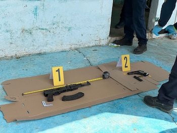 Un fusil, una pistola, municiones y herramientas de medición sobre cartón en un piso azul, marcados como evidencia. Se ven los pies de agentes y una pared clara