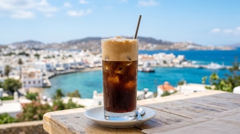 Un vaso de café frío con hielo, espuma y una pajita metálica sobre un plato blanco en una mesa de madera frente al mar.