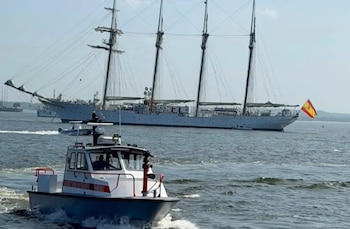 Una lancha blanca y roja en primer plano sobre el agua; al fondo, un velero blanco de cuatro mástiles con una bandera de España en la popa