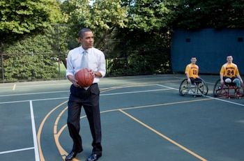 Barack Obama de pie en una cancha verde, sosteniendo un balón de baloncesto. Dos hombres en sillas de ruedas con camisetas amarillas de "Marines" están detrás de él