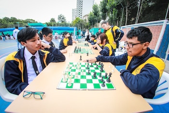 Jóvenes en uniformes azules y amarillos juegan ajedrez en mesas al aire libre; en primer plano, dos chicos se miran mientras mueven piezas en un tablero