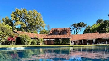 Piscina de agua azul en primer plano frente a una casa con tejado de tejas rojas y vegetación de fondo