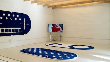 Un espacio con paredes blancas, tres alfombras ovoides de color azul y blanco en el suelo y la pared, y un televisor sobre un soporte metálico
