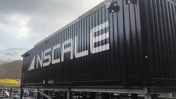 Primer plano de un gran contenedor negro con el logo blanco 'NSCALE' en su lateral, situado al aire libre con montañas nevadas y cielo nublado de fondo