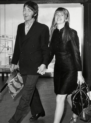 Fotografía en blanco y negro de Paul McCartney y Jane Asher caminando tomados de la mano y portando bolsos en un espacio interior