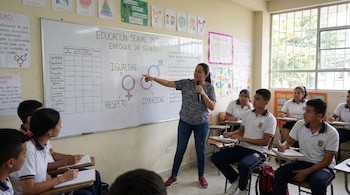 Una profesora señala una pizarra con el título Educación Sexual Integral Enfoque de Género a varios estudiantes uniformados sentados en pupitres en un aula.