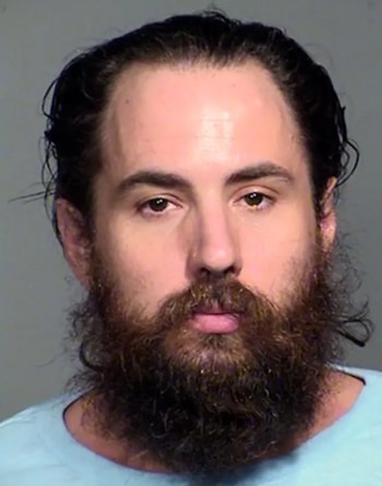 Retrato policial de un hombre de cabello oscuro y barba densa que viste una camiseta azul claro sobre un fondo gris