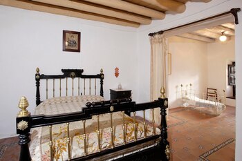 Dormitorio con cama de hierro y latón negro, mesita de noche, cuadro en la pared, vigas de madera en el techo, suelo de baldosas y una cuna en segundo plano